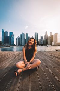 Portrait of smiling young woman sitting on cityscape against sky