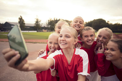 Cheerful soccer girls making faces while taking selfie from mobile phone against sky