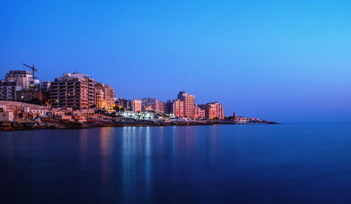 Illuminated buildings by sea against clear sky at dusk