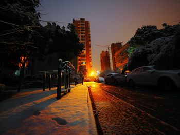 Snowy sidewalk by cars against sky during sunset