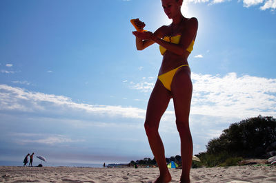 Young woman in bikini standing at beach against sky