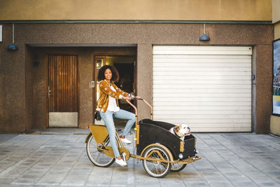 Full length of mid adult woman riding bicycle cart with dog against building