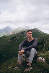 Portrait of young man sitting on mountain against sky
