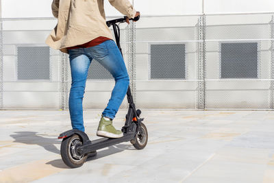 Back view of faceless ethnic male standing on modern electric scooter near metal wall on sunny day in city