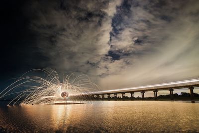 Person spinning wire wool against illuminated bridge over river at night