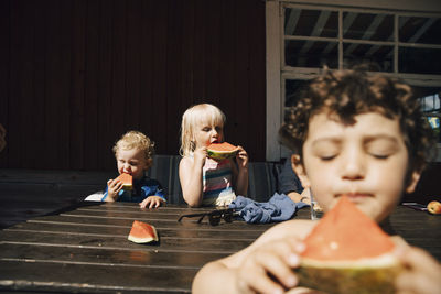 Siblings eating fruit while standing by table during sunny day