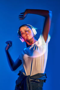Charming brazilian female listening to music in wireless headphones while dancing on purple background in studio with shadows on disco ball and fluorescent lights