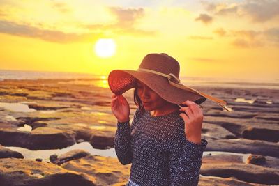 Young woman wearing hat standing at beach against sky during sunset