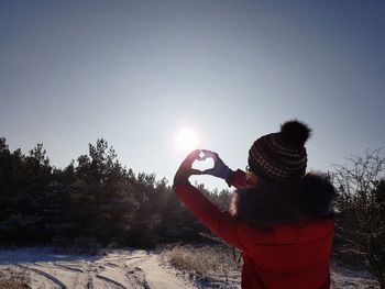 Rear view of woman making heart shape with hand against sky during winter