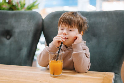 Four year old boy drinks orange juice from a straw while sitting at a table in a cafe and closes