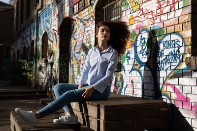 Portrait of confident young woman sitting on wood against graffiti wall in city