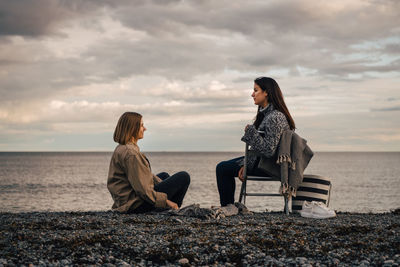 Female friends talking while sitting at beach against cloudy sky