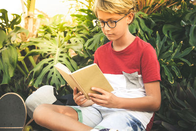 Boy reading book while sitting against plants