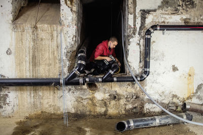 Male plumber connecting pipes amidst walls in basement