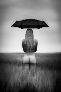 Rear view of naked woman holding umbrella while standing on field against sky