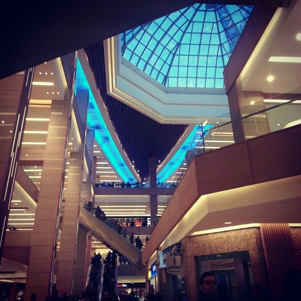 indoors, illuminated, architecture, built structure, low angle view, ceiling, modern, lighting equipment, blue, railing, transportation, incidental people, night, escalator, travel, shopping mall, architectural column, multi colored, interior, subway station