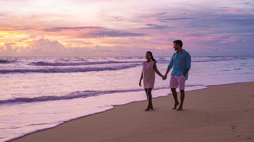 Rear view of couple walking at beach against sky during sunset