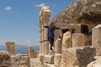 Young woman with arms outstretched walking on rocks against sky during sunny day