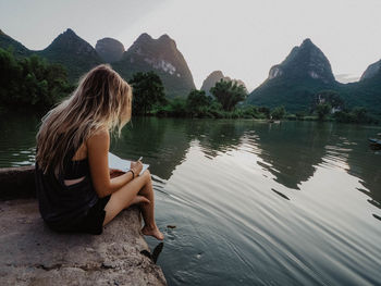 Woman sitting on rock by lake against clear sky