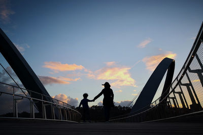 Silhouette mother holding hands of son while walking on bridge during sunset