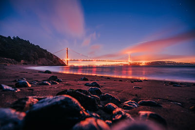 Illuminated suspension bridge over sea against sky during sunset