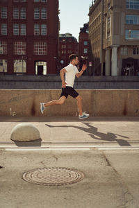Side view of strong male athlete jumping along street during active workout in city in summer