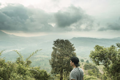 Side view of man looking at landscape while standing on mountain against cloudy sky