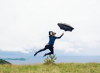 Young man jumping with umbrella on mountain against sea