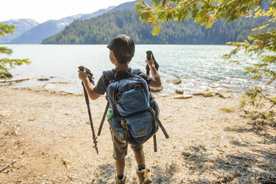 Rear view of young boy hiking by lake with backpack, hiking poles