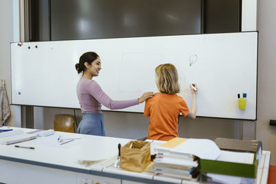 Smiling female student with hand on shoulder of student writing on whiteboard in classroom