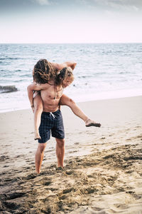 Full length of shirtless man giving piggyback to woman on shore at beach