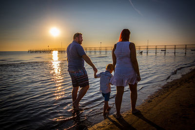 Rear view of parents holding son hands while walking on shore at beach against sky during sunset