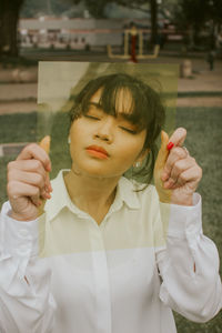 Young woman with eyes closed holding plastic in front of face while sitting outdoors