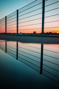 Reflection of fence in pond against sky during sunset