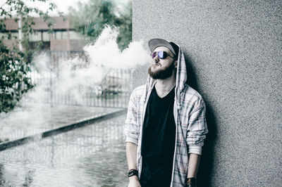 Young man smoking while standing by wall during rain