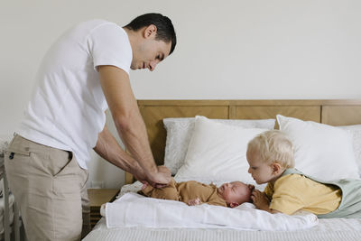 Boy looking at father buttoning baby's rompers on bed