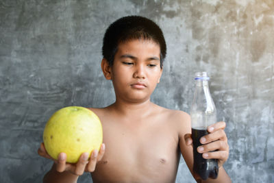 Shirtless boy holding food and drink while sitting against wall