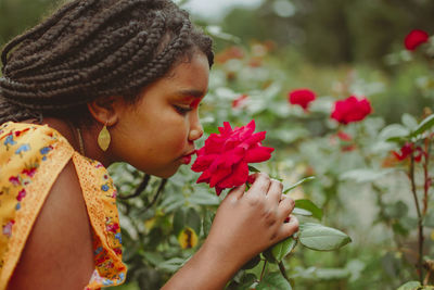 Side view of girl smelling red flower while standing in park