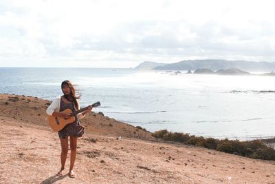 Woman with guitar standing on beach against sky