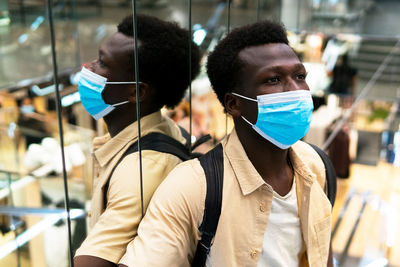 Calm african american male customer in medical mask standing near mirrored wall in modern shopping center and looking away during coronavirus