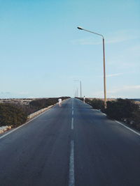 Empty road along street lights against clear blue sky
