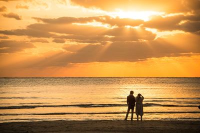 Rear view of man and woman standing at beach against cloudy sky during sunset