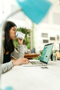 Side view of female buyer drinking beverage while sitting at table with laptop and choosing goods during online shopping