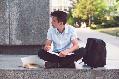 Young man studying while sitting by wall