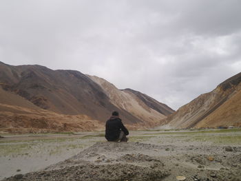 Rear view of man relaxing by lake against cloudy sky over mountain range