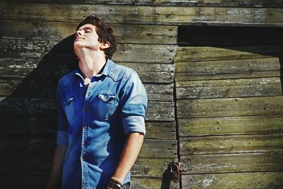 Young man leaning on weathered wooden wall during sunny day