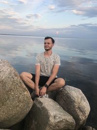 Young man sitting on rock by sea against sky