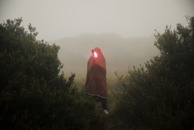 Man with illuminated face covered in blanket while standing amidst plants on mountain