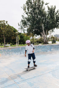 Full body of young ethnic person in casual outfit wearing protective helmet with knee pads and elbow pads riding skateboard in skate park