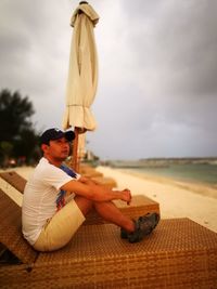 Mid adult man sitting on deck chair at beach against sky